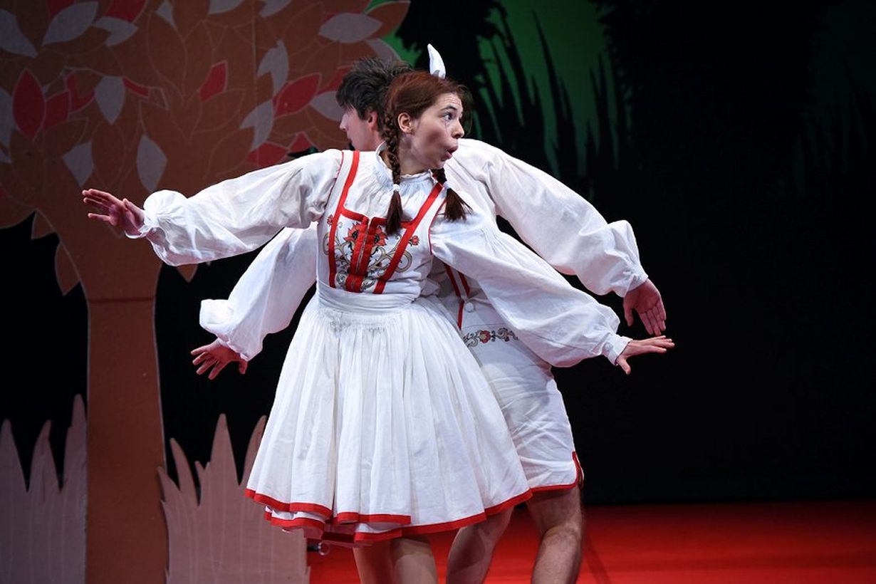 Two actors in a play, portraying Janko and Metka. They are dressed in white outfits and are positioned back to back.